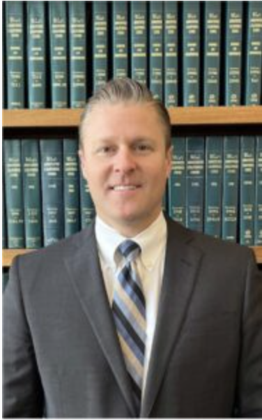 White male in a suit and tie standing in front of law books on shelves. 